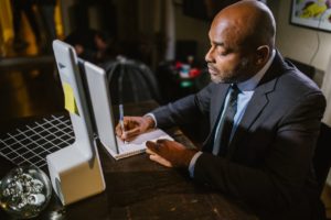 black man writing in notebook with tablet screen in front of him