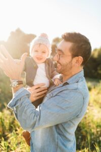 Man in field holding a baby. Both are smiling. No one knows why.