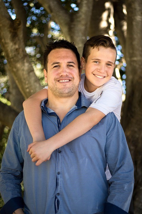 A man being hugged around the neck from behind by a child who somewhat resembles him. There is a tree behind them. The tree is just standing there.